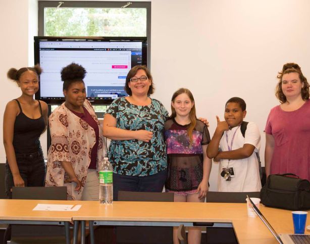 Copyright 2024 Digital Bridges International. All rights reserved. Founder Connie Capiotis. Group of smiling diverse students in a classroom posing with Connie.